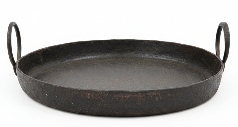 A round, dark gray cast iron serving tray with two loop handles, resting on a rustic wooden cabinet inside a home decor store.