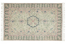 A rectangular vintage-style area rug with fringe on the sides, featuring a faded central medallion and geometric borders in muted pink, sage green, and gray on a light beige ground, laid flat on a dark gray floor.