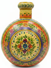 A round hand-painted wooden decorative vase with a narrow neck, featuring intricate multicolor floral and leaf patterns on a yellow background, set on a dark wooden table in a shop interior.