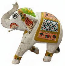 A hand-painted wooden elephant figurine with a distressed white finish, decorated with vibrant orange, green, yellow, and blue patterns, standing on a dark wooden table in a store setting.