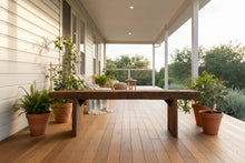A rustic dark brown wooden coffee table with thick block legs, shown in a showroom setting with a distressed gray floor and other furniture in the background.