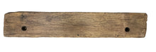 A long, rustic wooden plank or bench with two circular holes, photographed from above on a gray concrete floor next to a crate. A person's feet and a colorful bag are partially visible at the edges.