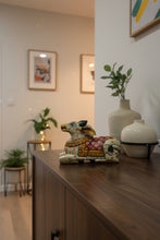 A hand-painted timber figurine of a cow lying down, decorated with traditional patterns in red, yellow, green, and black on a light beige base, set on a wooden table in a shop.