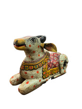A hand-painted wooden cow figurine with traditional Indian motifs, resting on a dark wooden table in a shop setting.