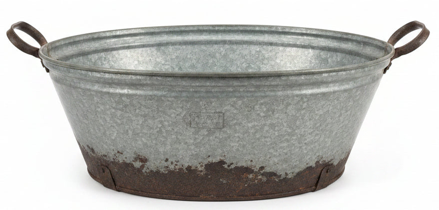 A large galvanized metal tub with side handles, showing a distressed and rusted bottom, sitting on a dark wooden table in a home decor store.