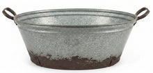 A large galvanized metal tub with side handles, showing a distressed and rusted bottom, sitting on a dark wooden table in a home decor store.