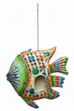 A colorful hand-painted fish-shaped wall hanging or decorative piece suspended in front of an ornate carved wooden doorway. The fish design features multiple bright colors including green, blue, orange, pink, and yellow, with a cutout in the center revealing a gray cat inside.