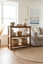 A rustic two-tier wooden side table or shelf made from natural wood, set against a background of other antique furniture and a colorful painted wooden panel.