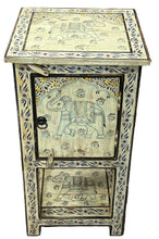A small carved wooden cabinet with a single door and a lower shelf, featuring hand-painted elephant motifs and dark floral border details, photographed on a gray floor in a rustic indoor setting.