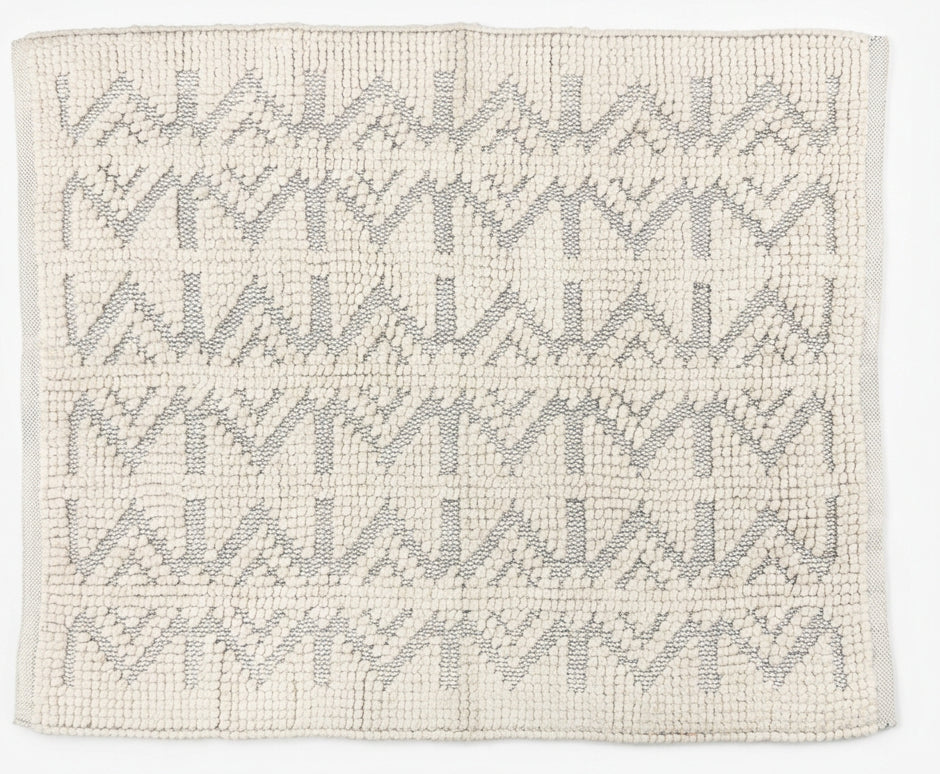 A rectangular woven rug with a gray geometric pattern on an off-white base, laid out on a dark gray concrete floor. The bottom of the frame shows a person's feet wearing dark brown shoes and dark pants, and a black-and-white decorative object on the right.