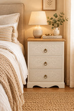 A three-drawer chest of drawers in a distressed white finish with carved floral patterns on the drawer fronts and a natural brown wood top, set against a plain white wall and dark floor.